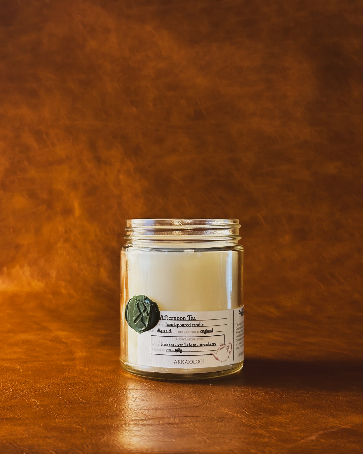 Afternoon Tea candle jar with green wax seal and white label inspired by artifact tag, no lid on leather background