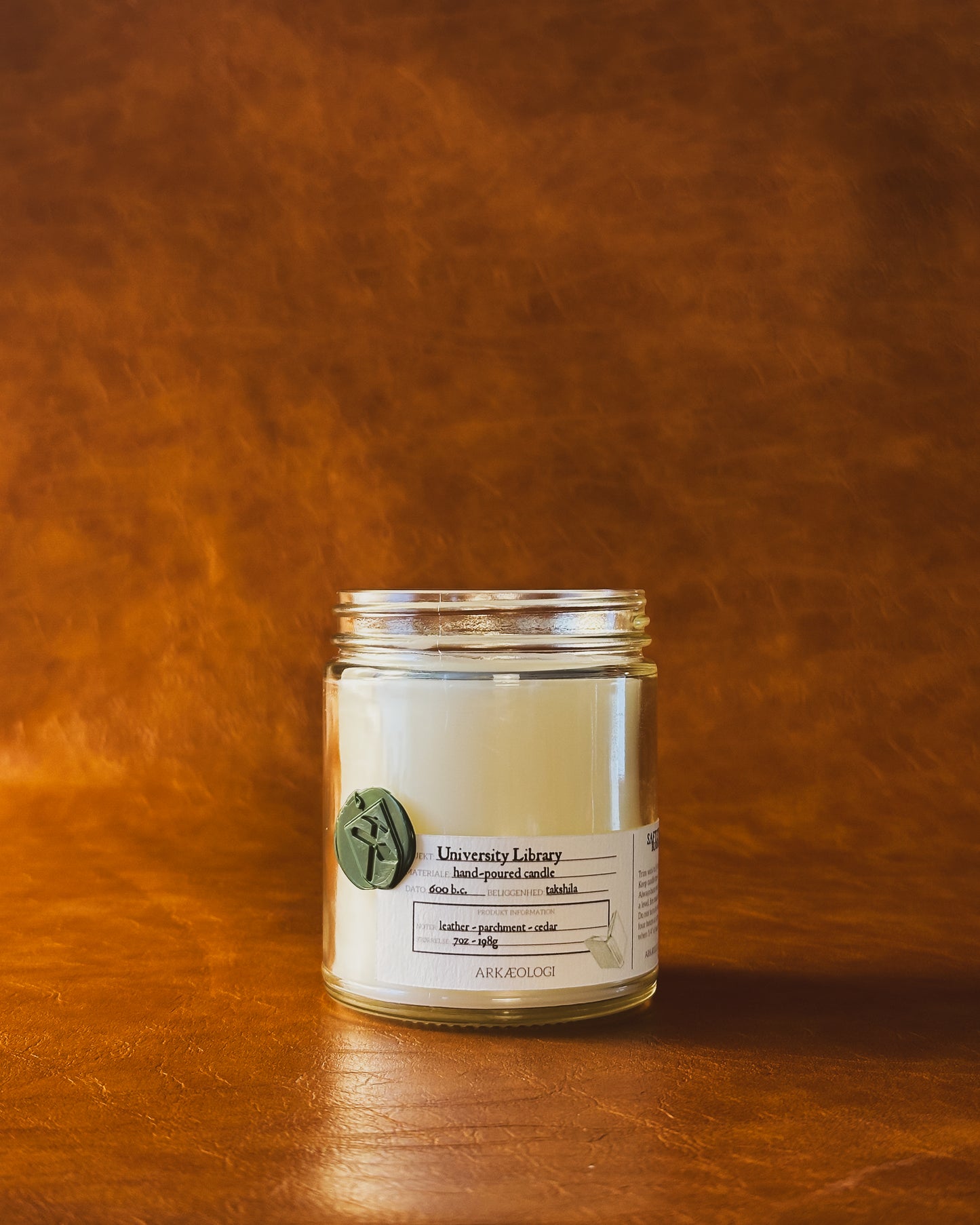 University Library candle jar with green wax seal and white label inspired by artifact tag, no lid on leather background