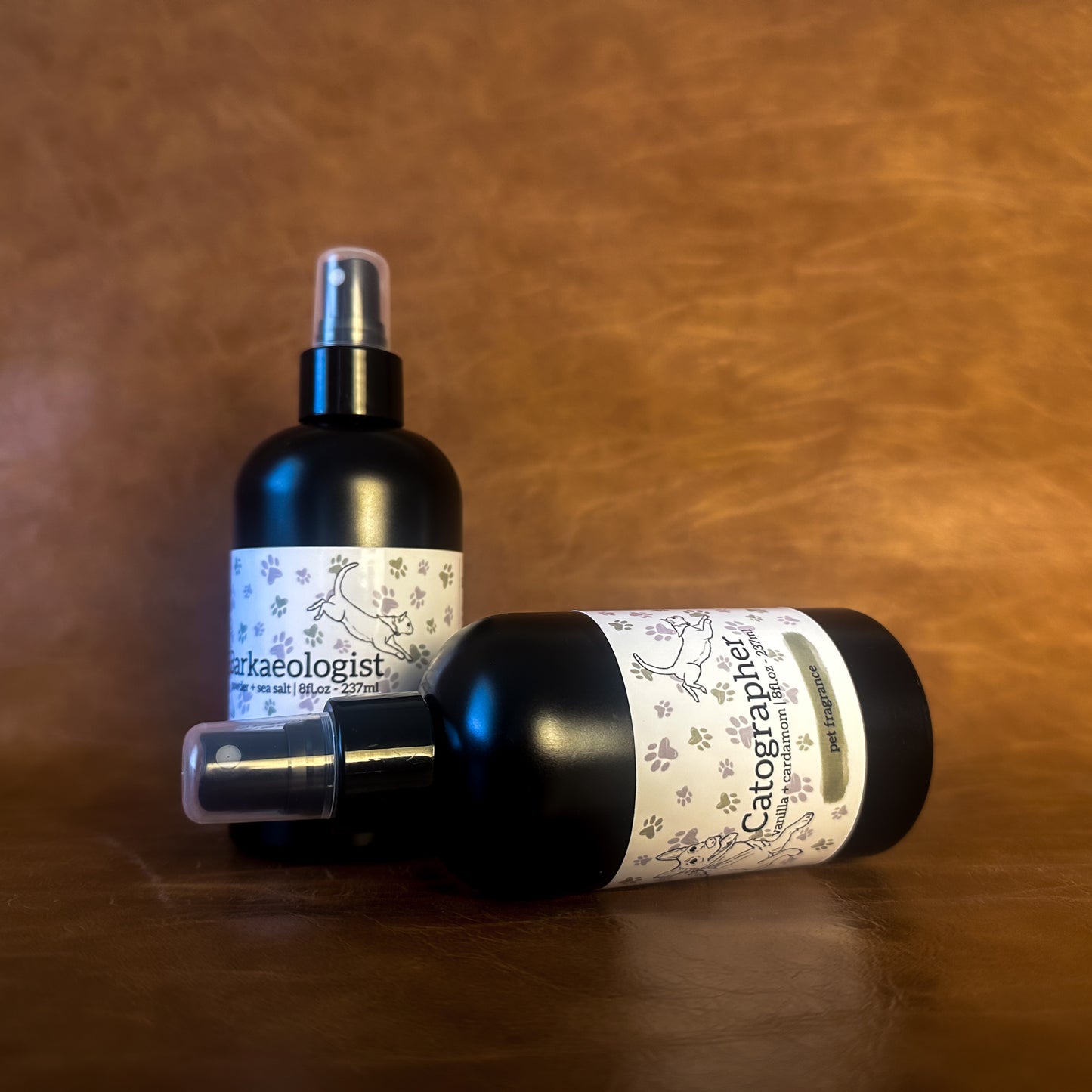 Two black pet perfume bottles with a white label featuring an illustration of a dog and cat on a brown leather background.