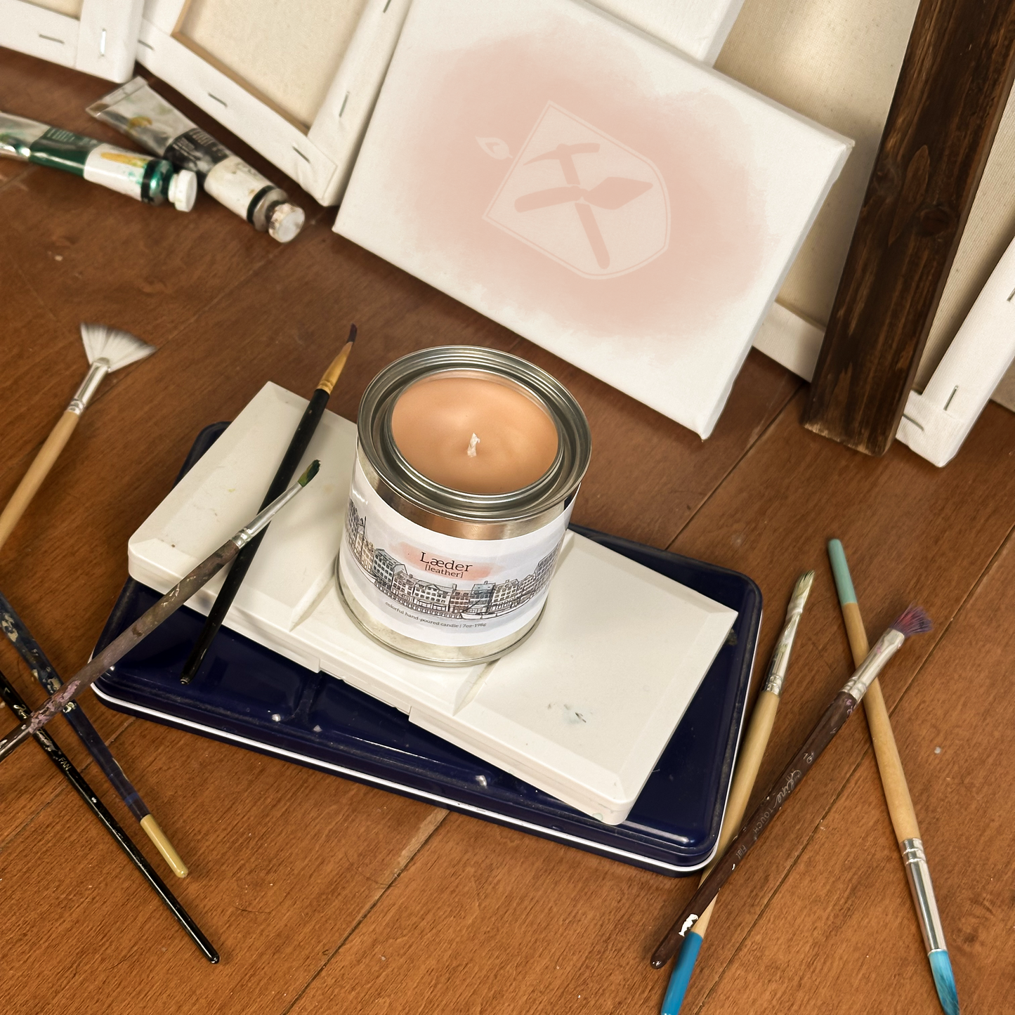 Orange candle in paint can surrounded by paint supplies on a wood floor. The Label on the candle depicts the Nyhavn and reads "Leather" in Danish