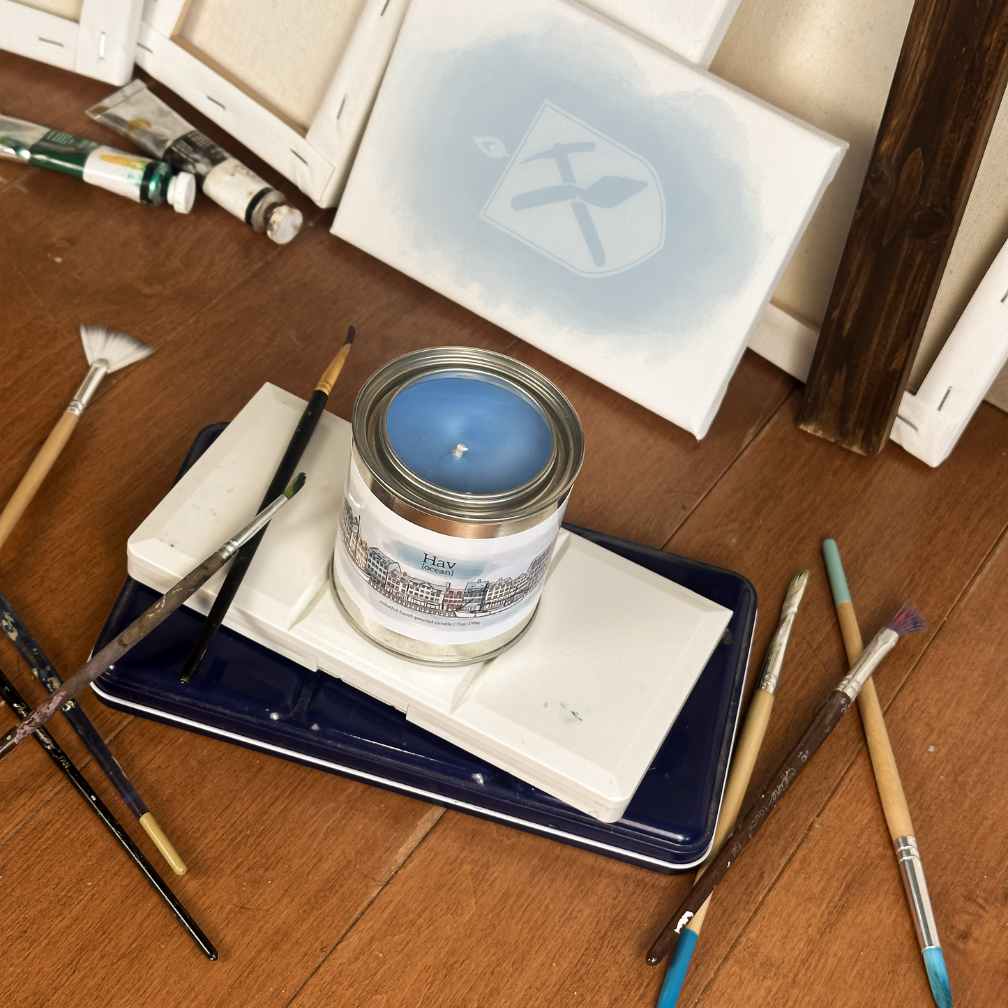 Blue candle in paint can surrounded by paint supplies on a wood floor. The Label on the candle depicts the Nyhavn and reads "Ocean" in Danish