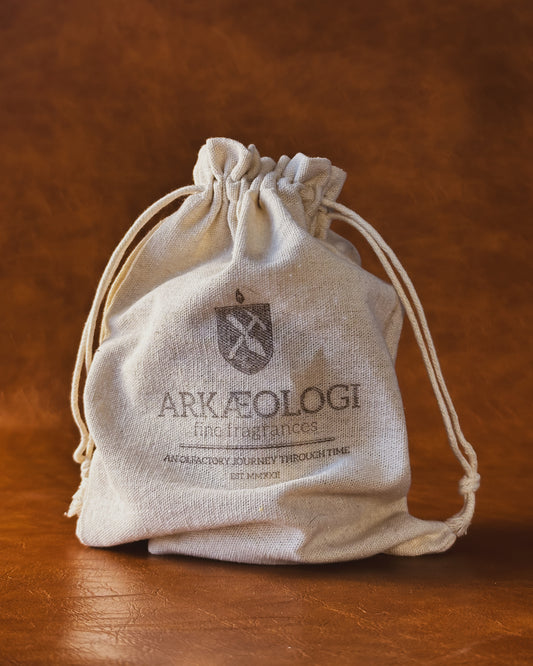 Beige drawstring bag on a brown leather background. The bag has printed "Arkæologi, fine fragrances, an olfactory journey through time, est. MMXXII"