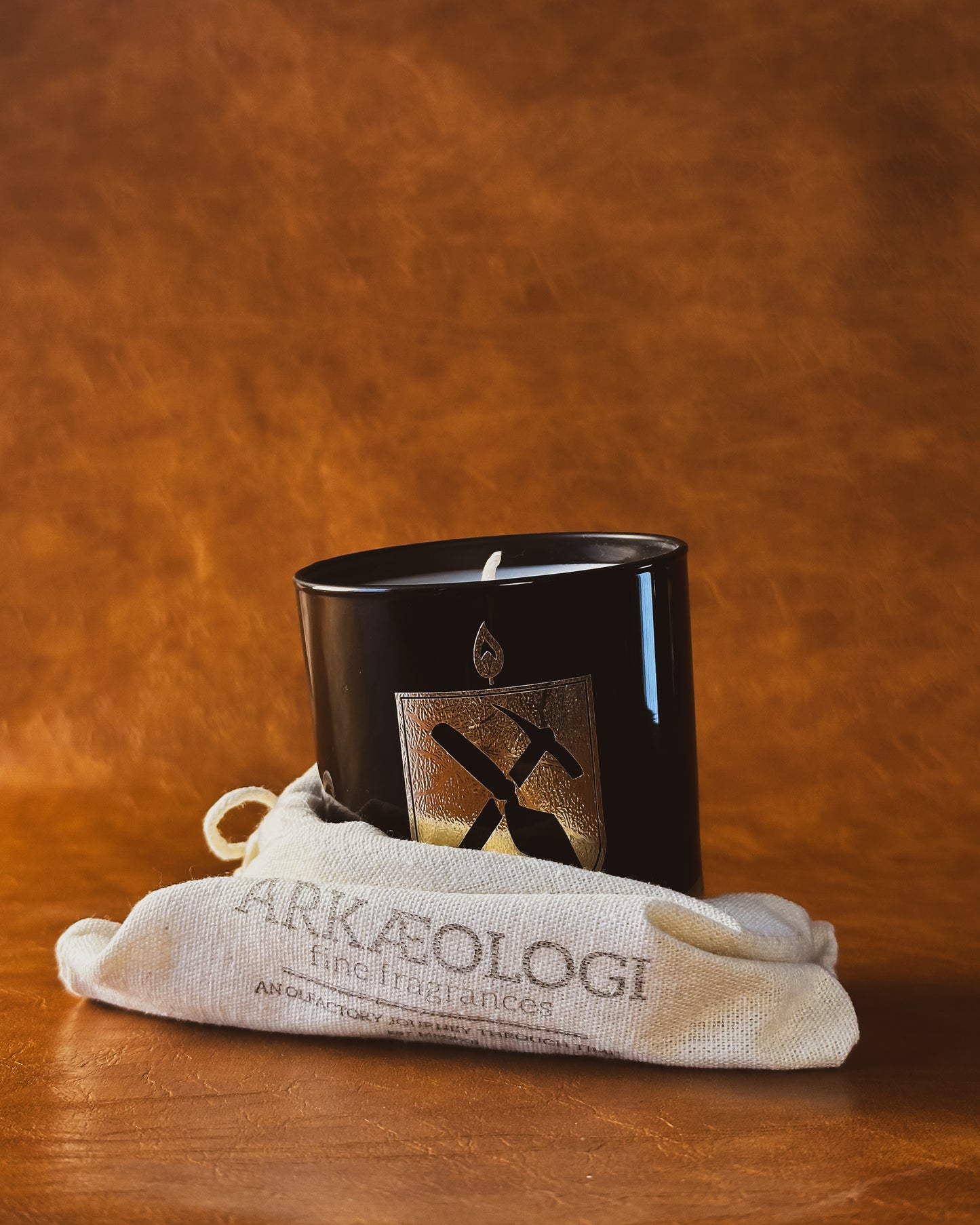 Black glass candle with gold Arkæologi crest in a beige drawstring bag on a brown leather background. The bag has printed "Arkæologi, fine fragrances, an olfactory journey through time, est. MMXXII"