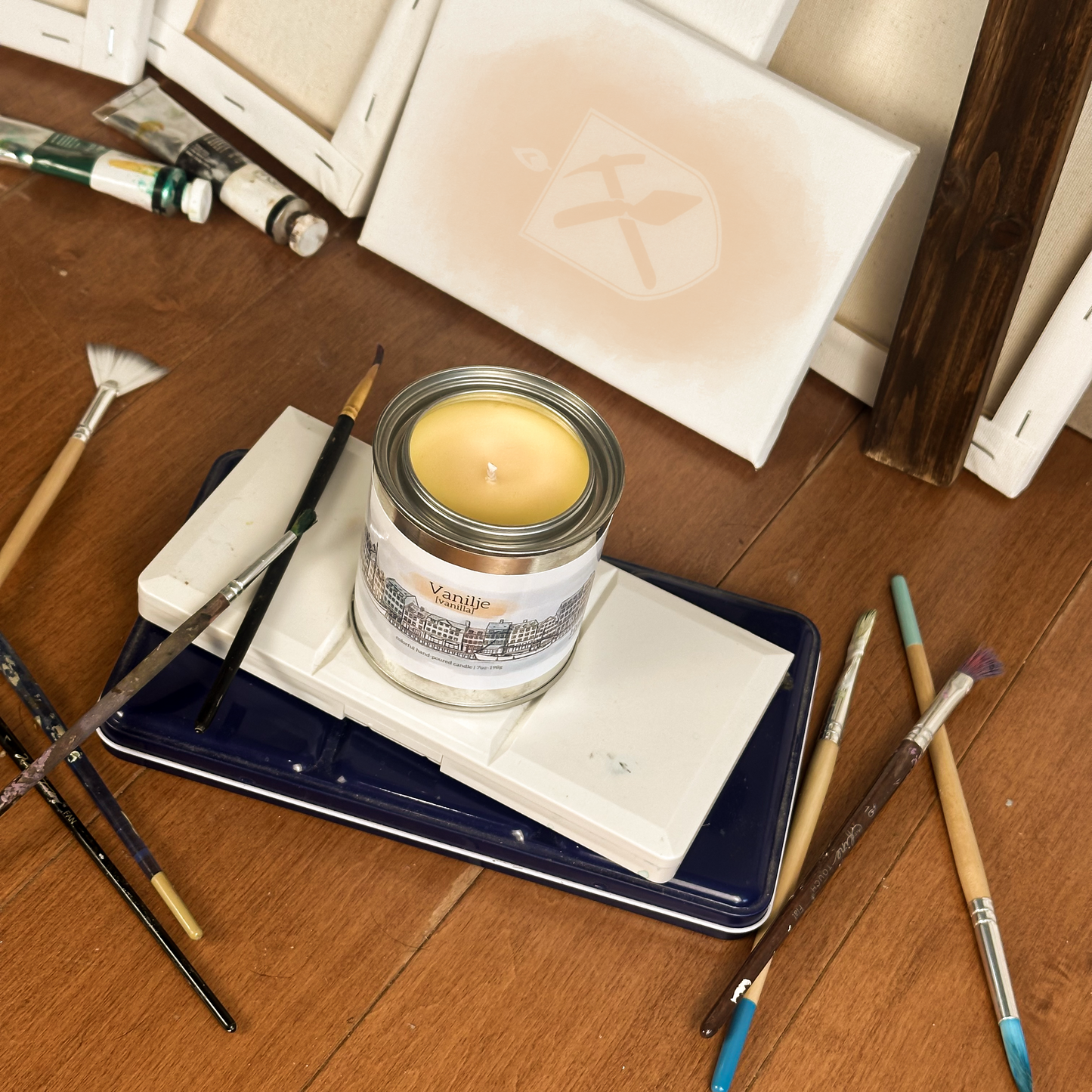Yellow candle in paint can surrounded by paint supplies on a wood floor. The Label on the candle depicts the Nyhavn and reads "Vanilla" in Danish