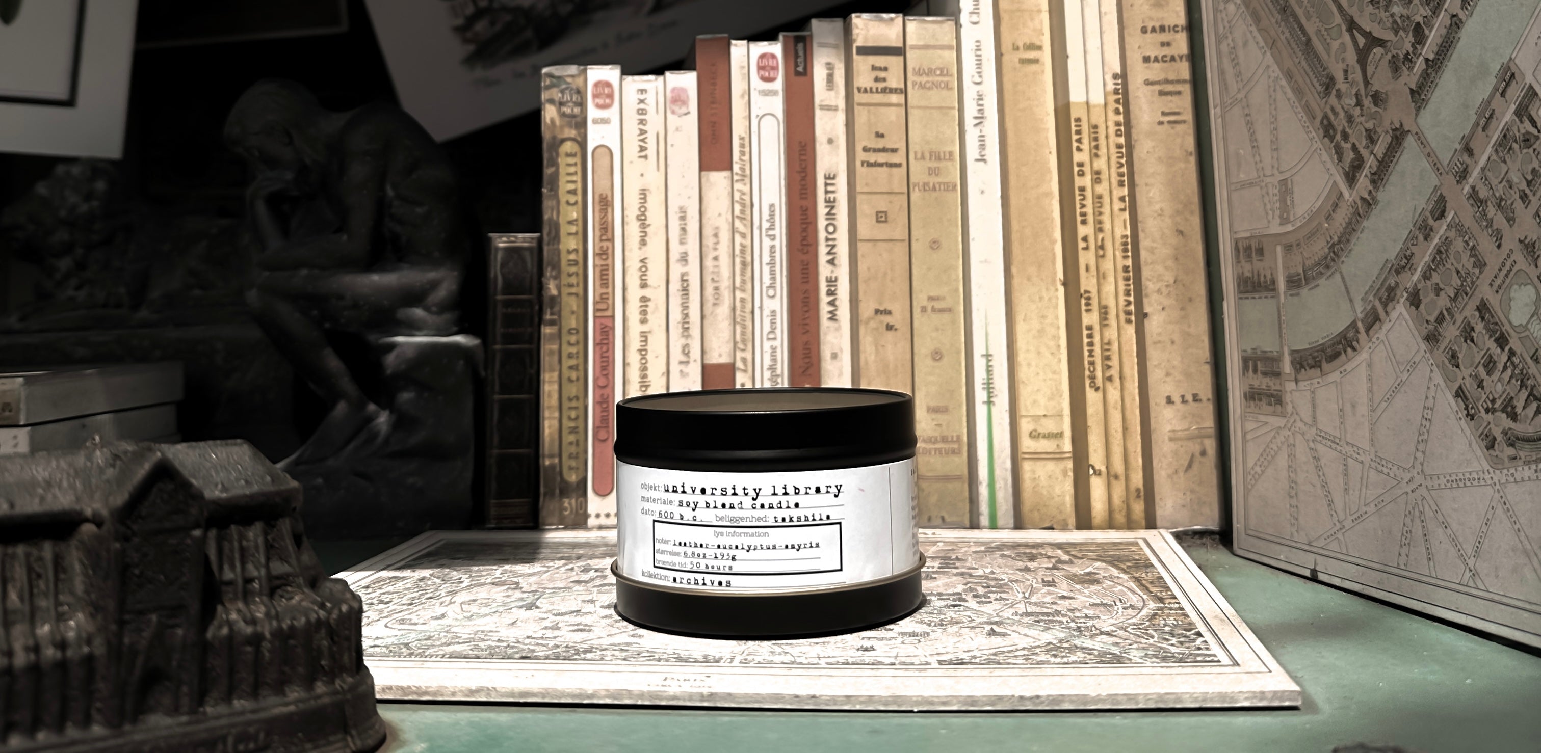 Arkaeologi University Library scented candle in a black tin jar on a green shelf with books and maps.