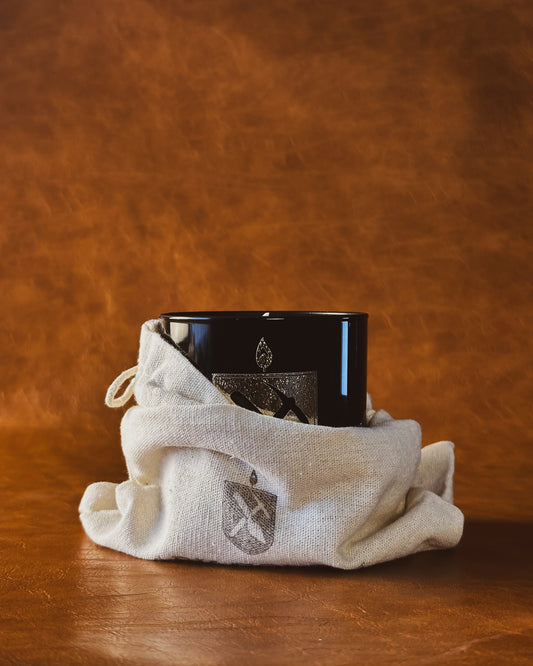 Black glass candle with gold Arkæologi crest in a beige drawstring bag on a brown leather background.