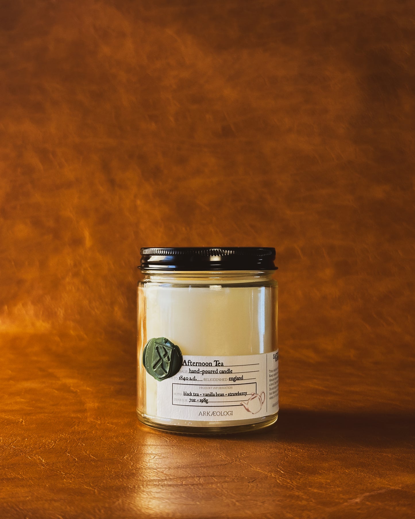 Afternoon Tea candle in clear jar with green wax seal and white label inspired by artifact tag, with black lid on leather background