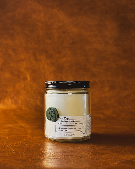Aqua Virgo candle in clear jar with green wax seal and white label inspired by artifact tag, with black lid on leather background
