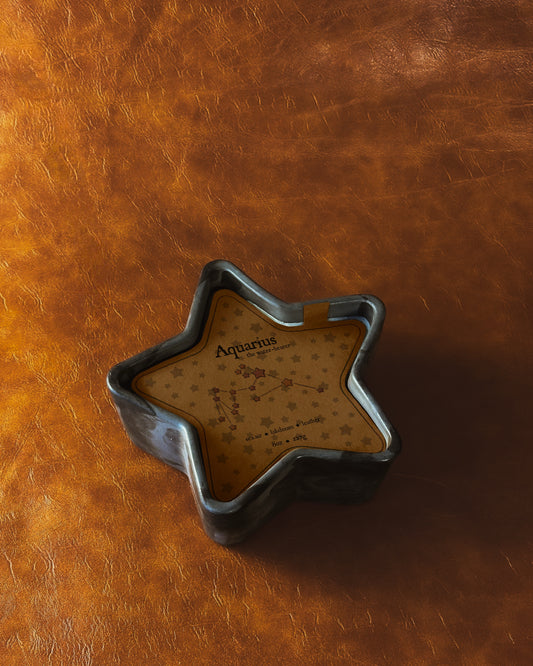 Black concrete star shaped candle with brown insert that reads "Aquarius" and has the constellation in purple and yellow on a brown leather background.