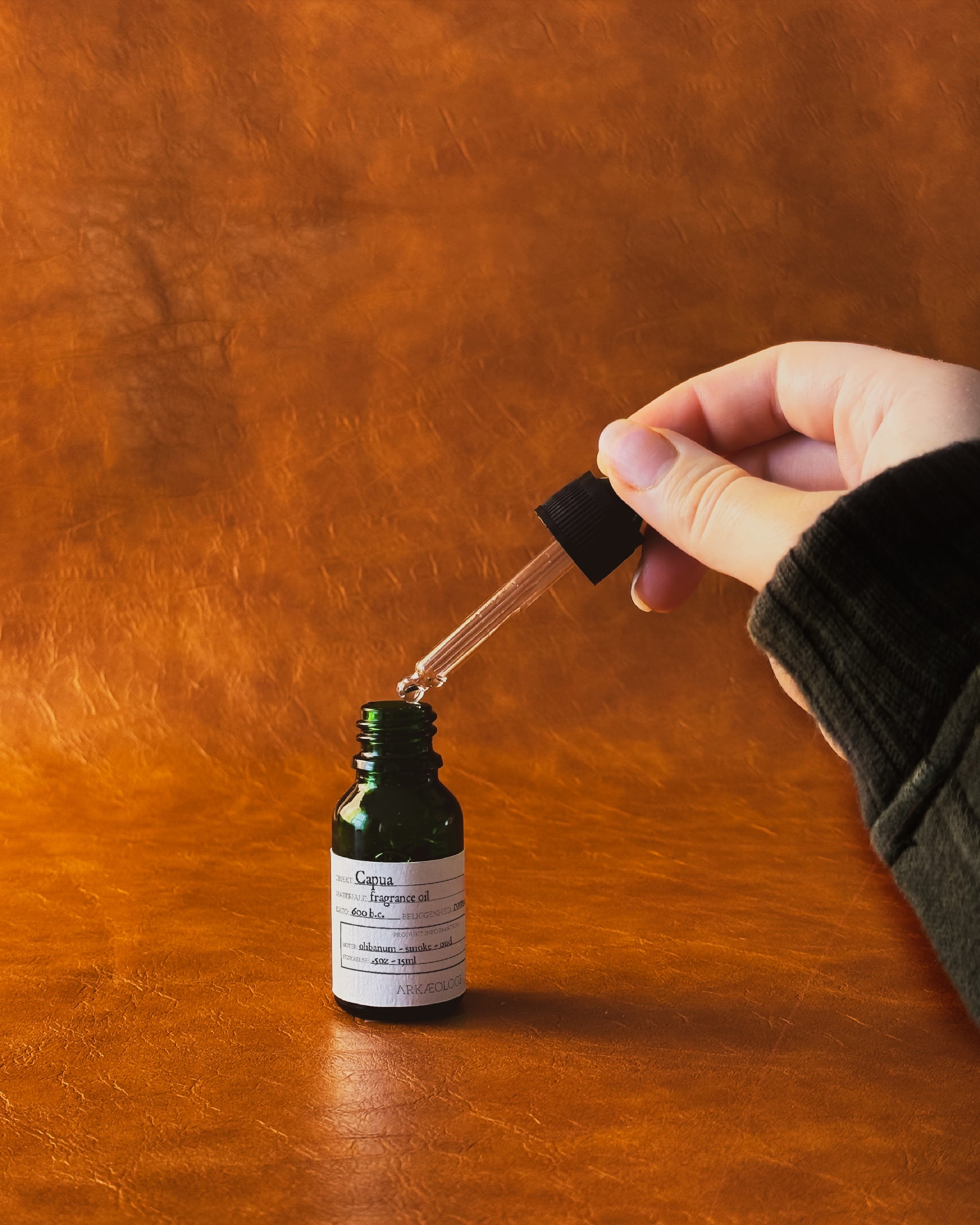 Hand holding a glass dropper over a green fragrance oil bottle labeled "Capua" on a leather background.