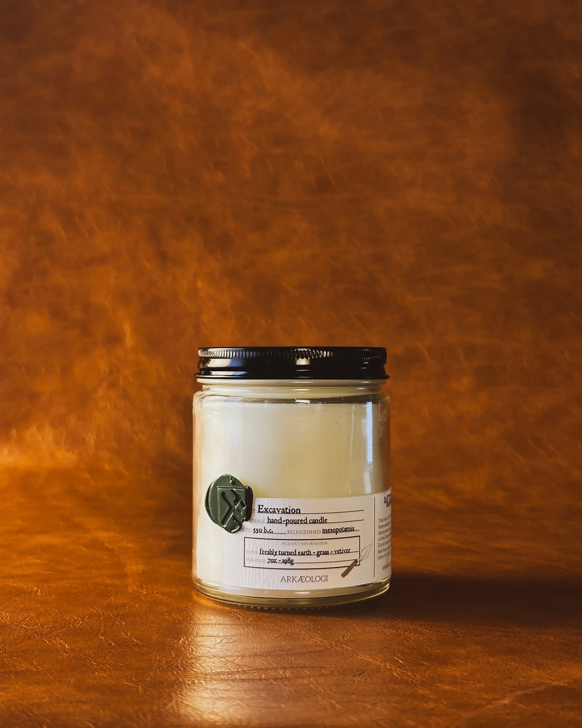 Excavation candle in clear jar with green wax seal and white label inspired by artifact tag, with black lid on leather background