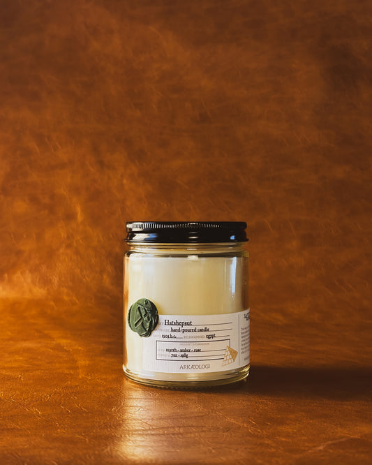 Hatshepsut candle in clear jar with green wax seal and white label inspired by artifact tag, with black lid on leather background