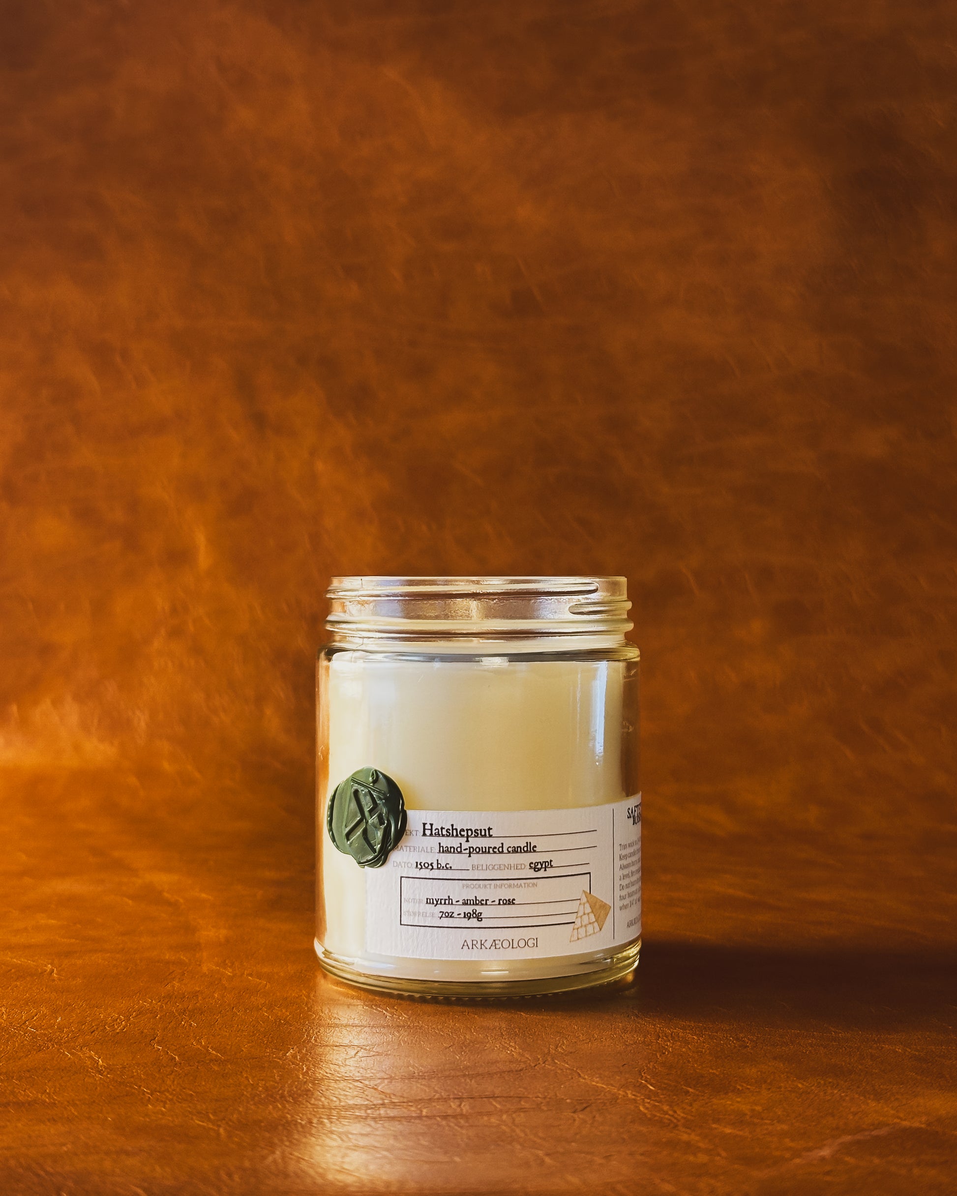Hatshepsut candle jar with green wax seal and white label inspired by artifact tag, no lid on leather background