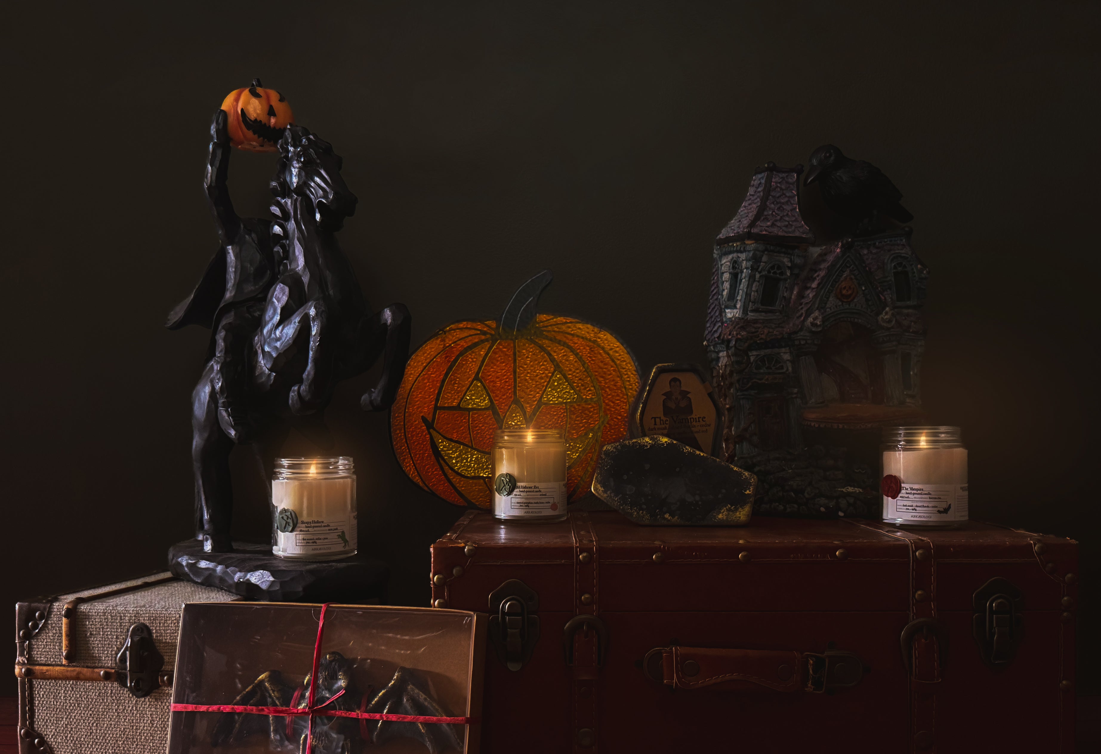Halloween decor on two tanks. A black headless horseman figurine holding a carved pumpkin, a orange jack-o'-lantern stained glass, a dark haunted-house decoration with a crow on top, three lit candles (sleepy hollow, all hallows' eve, and the vampire), a coffin candle with gold detailing, a benny the bat candle holder in a brown paper box all on a dark green background.