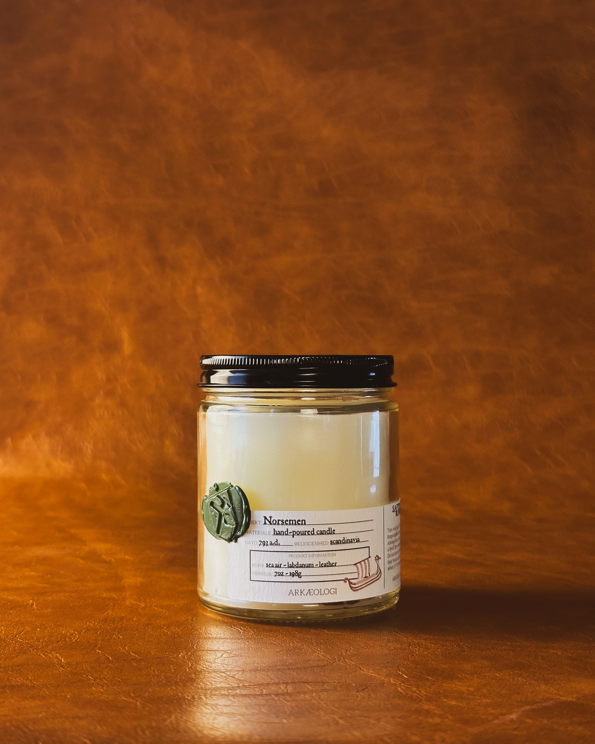 Norsemen candle in clear jar with green wax seal and white label inspired by artifact tag, with black lid on leather background