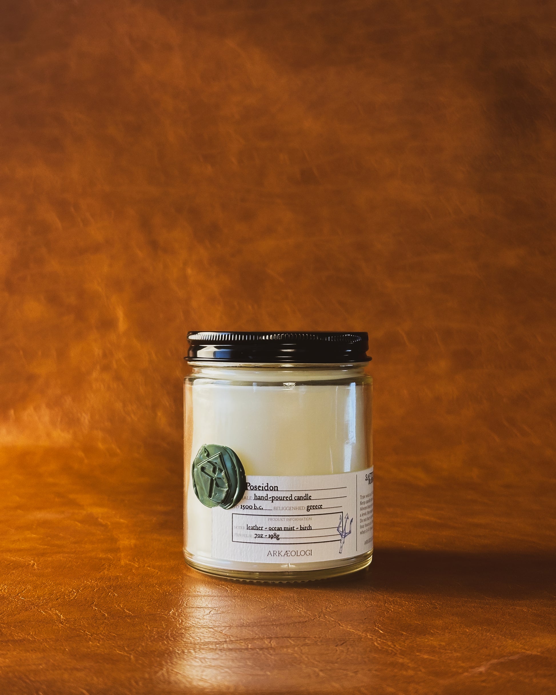 Poseidon candle in clear jar with green wax seal and white label inspired by artifact tag, with black lid on leather background