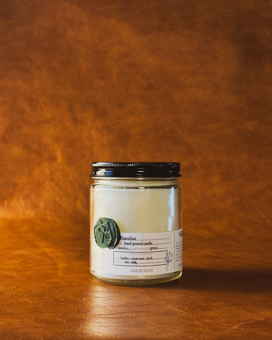 Poseidon candle in clear jar with green wax seal and white label inspired by artifact tag, with black lid on leather background