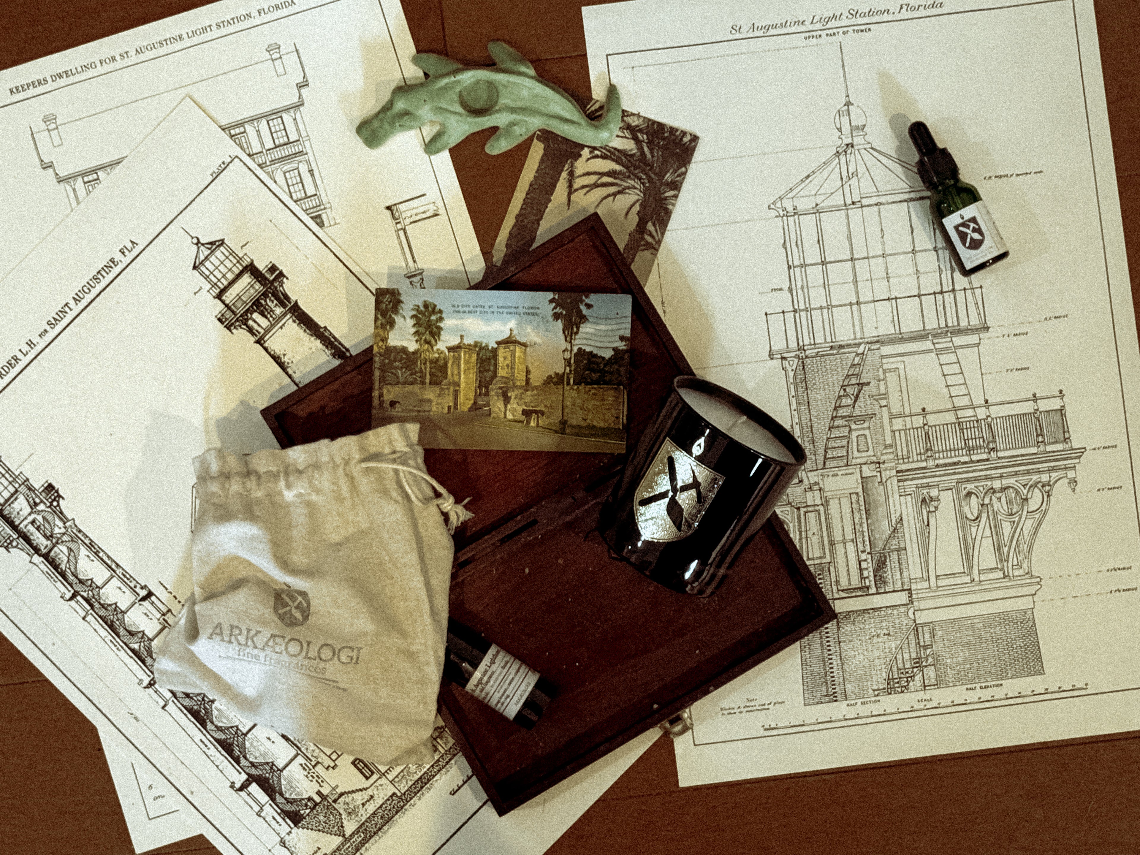 St. Augustine Lighthouse black glass candle and solid fragrance in open wood box with cream cloth bag, fragrance oil, postcards and prints of the St. Augustine Lighthouse on a wood background.