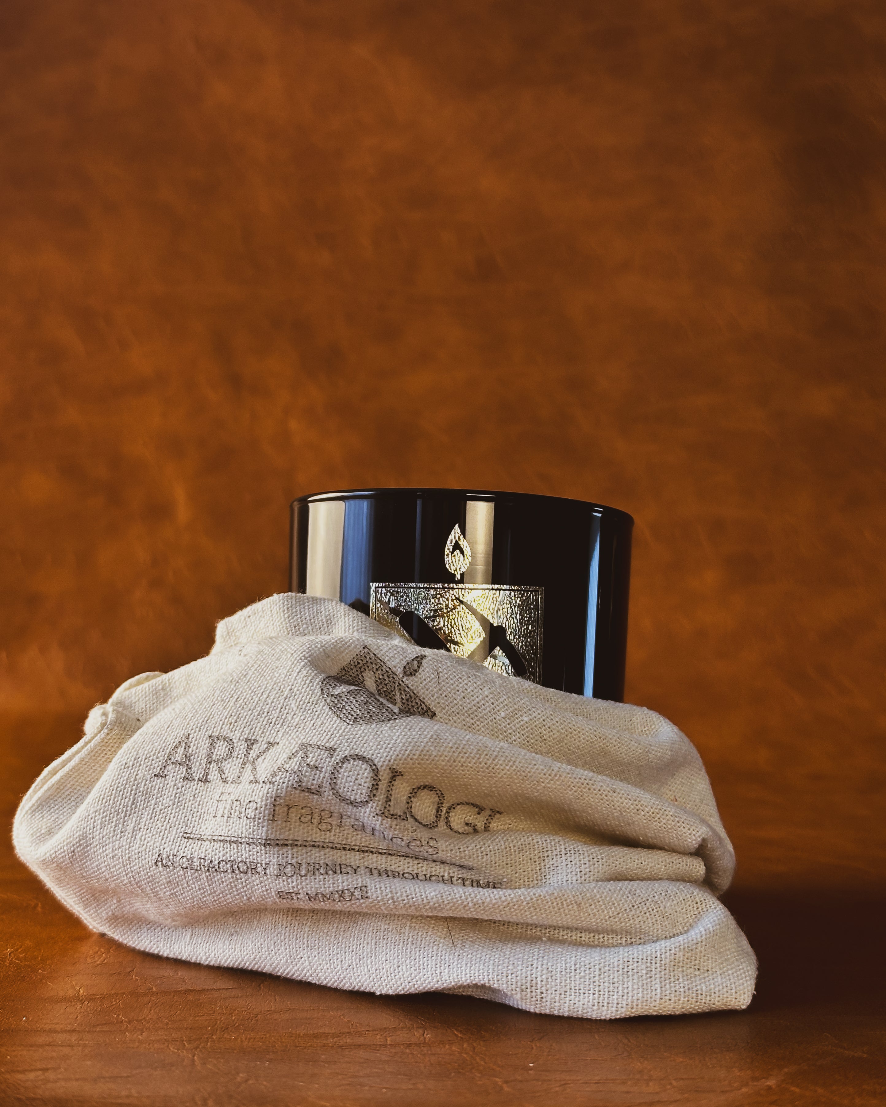 black glass candle with gold Arkæologi crest partly in cream cloth bag on leather background 