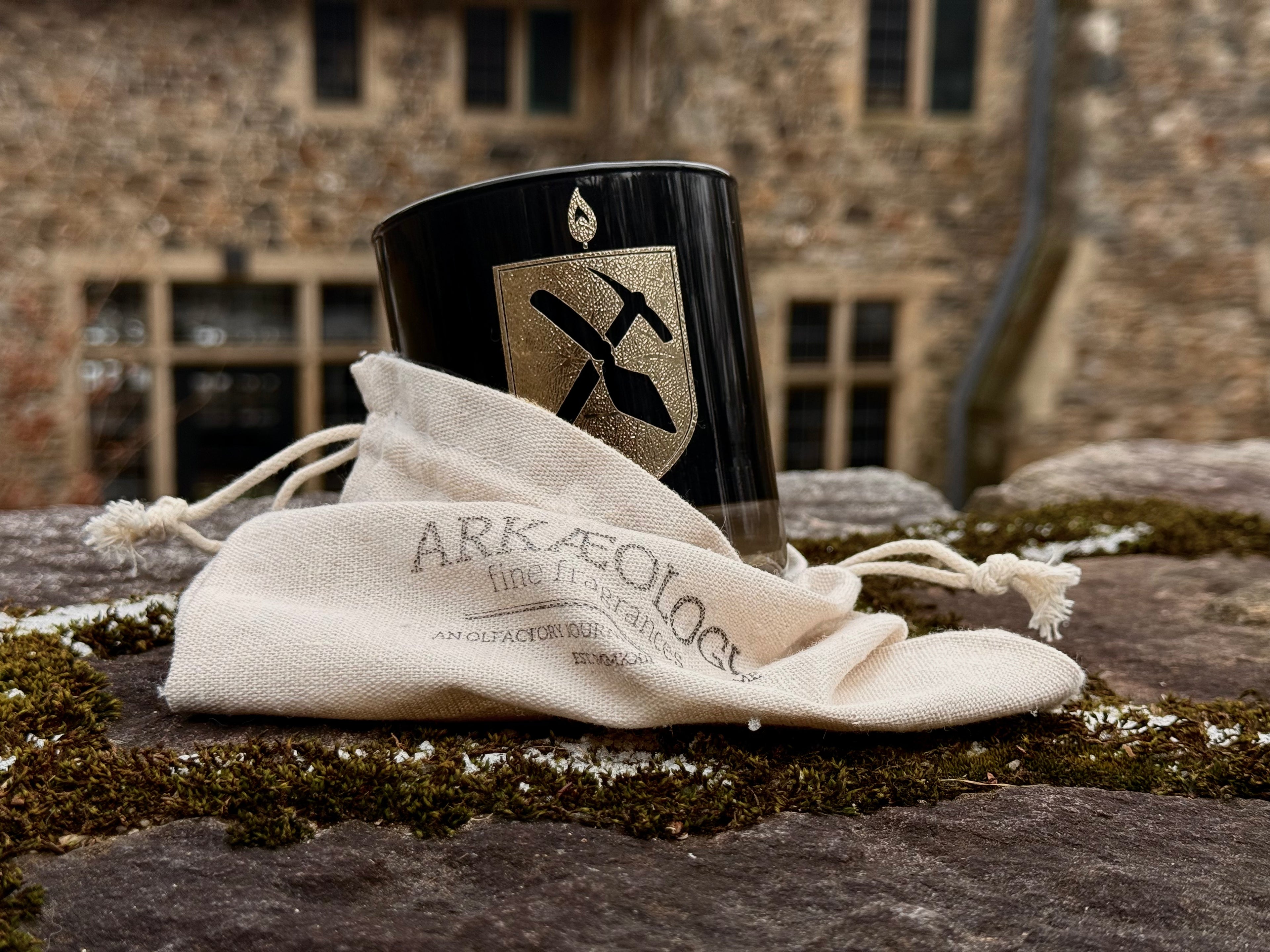 Candle in black rocks glass with gold Arkæologi logo, partially in a cream colored cloth bag with Arkæologi fine fragrances stamped on the front. The Candle is on a stone wall infront of an old stone building. 