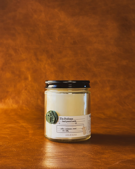 The Professor candle in clear jar with green wax seal and white label inspired by artifact tag, with black lid on leather background