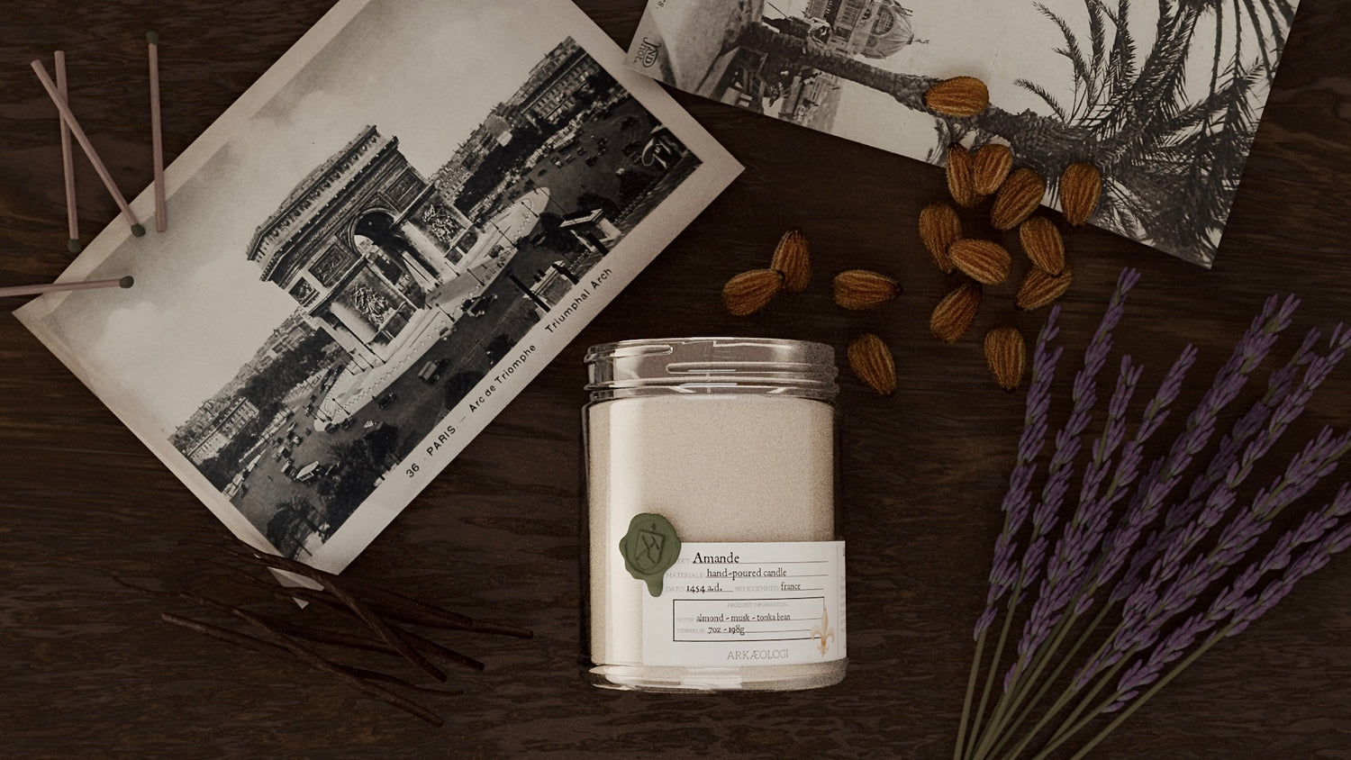 Amande Candle on wood table with French postcards, matches, almonds, dried lavender, and vanilla bean pods. 