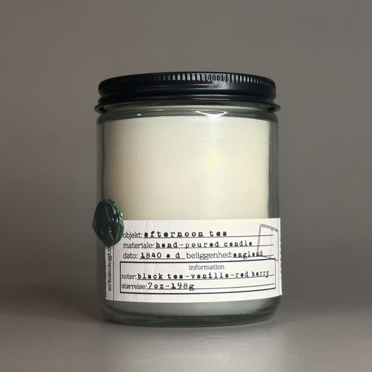 Glass jar candle with black lid and green wax seal of a trowel and pickaxe. Textured white label, themed after an artifact tag, reads "objekt: afternoon tea. materiale: hand-poured candle. dato: 1840 a.d. beliggenhed: England. noter: black tea-vanilla-red berry. størrelse: 7oz-198g." Candle is on a grey background.