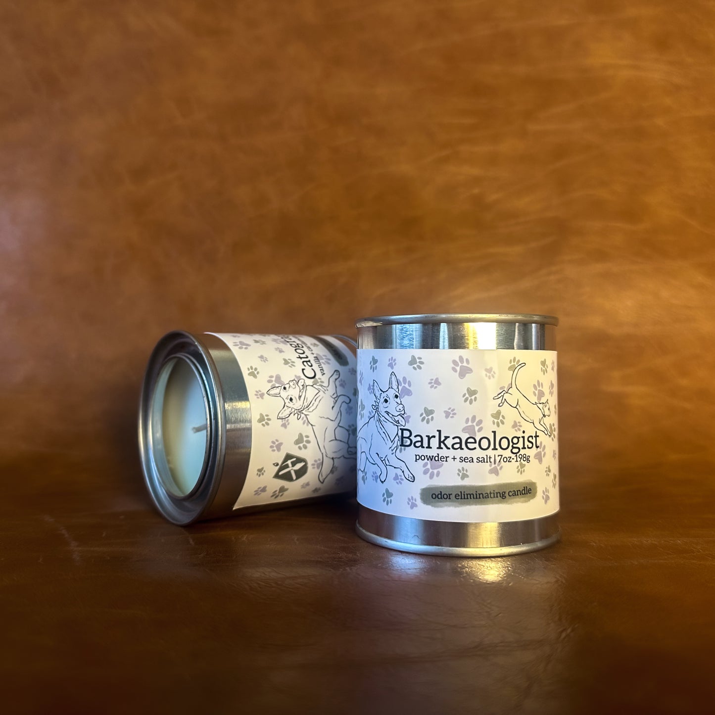 Two paint can candles with a white label featuring an illustration of a dog and cat on a brown leather background. The Label reads "Barkaeologist, powder + sea salt | 7oz-198g, odor eliminating candle"