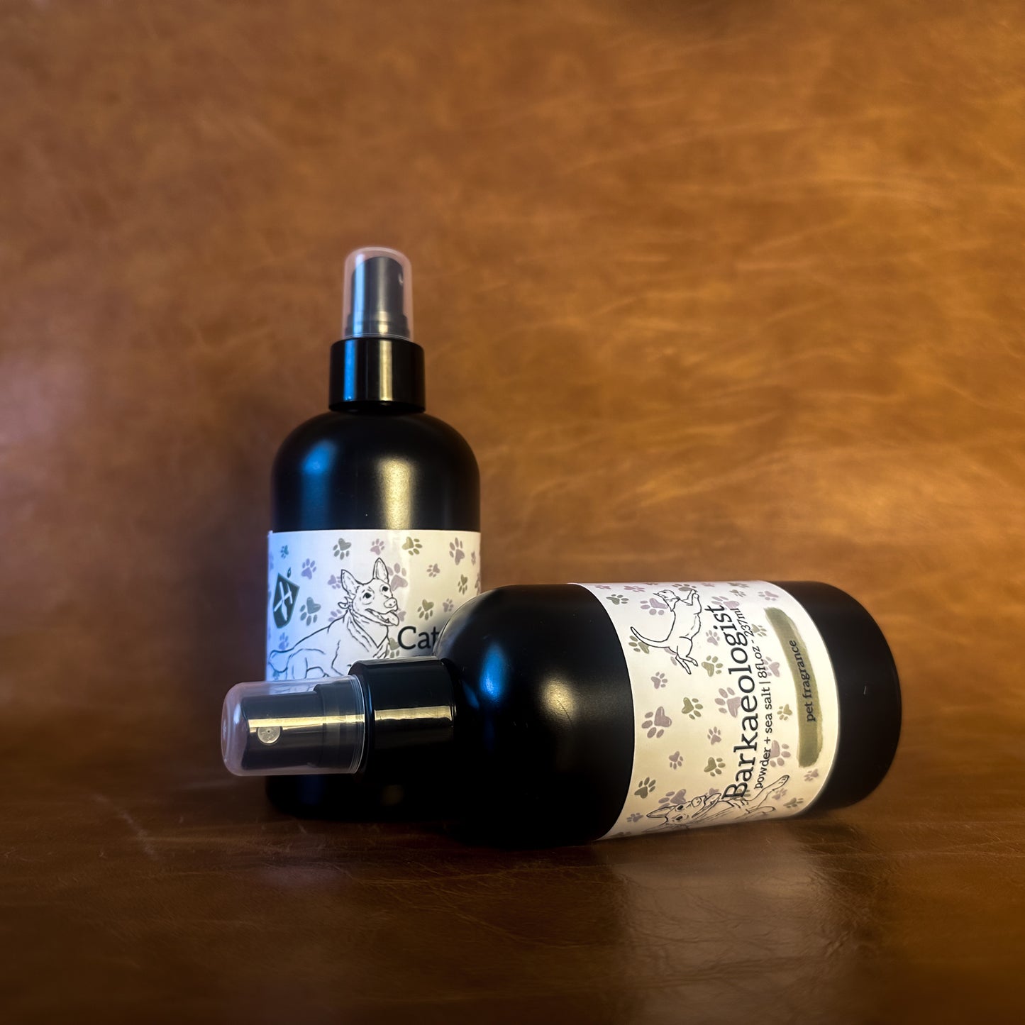 Two black pet perfume bottles with a white label featuring an illustration of a dog and cat on a brown leather background. 