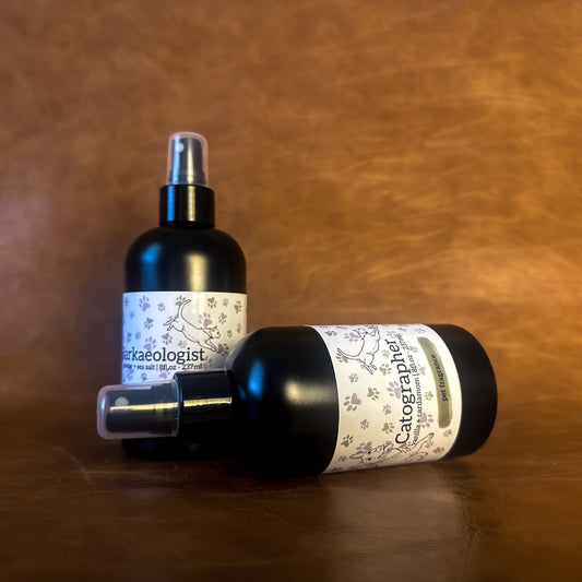 Two black pet perfume bottles with a white label featuring an illustration of a dog and cat on a brown leather background. 