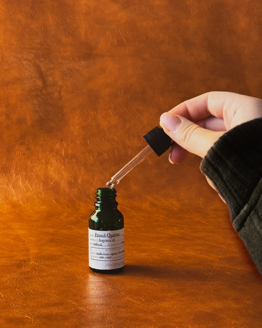 Hand holding a glass dropper over a green fragrance oil bottle labeled "French Quarter" on a leather background.